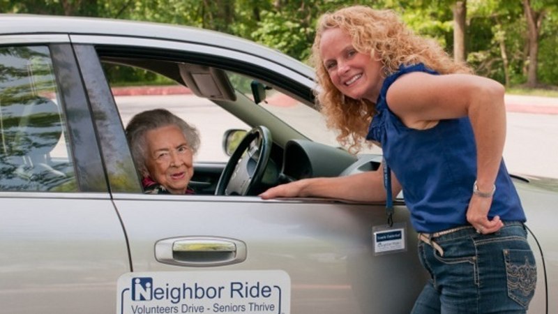 Neighbor Ride volunteer stands next to vehicle and smiles with older adult female who is sitting in the passenger seat