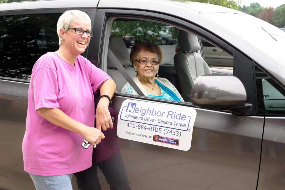 Neighbor Ride volunteer driver standing next to passenger side of vehicle with Neighbor Ride client sitting inside the car