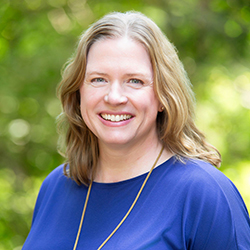 A woman with shoulder-length light brown hair, wearing a blue top and a necklace, smiles outdoors with green foliage in the background.