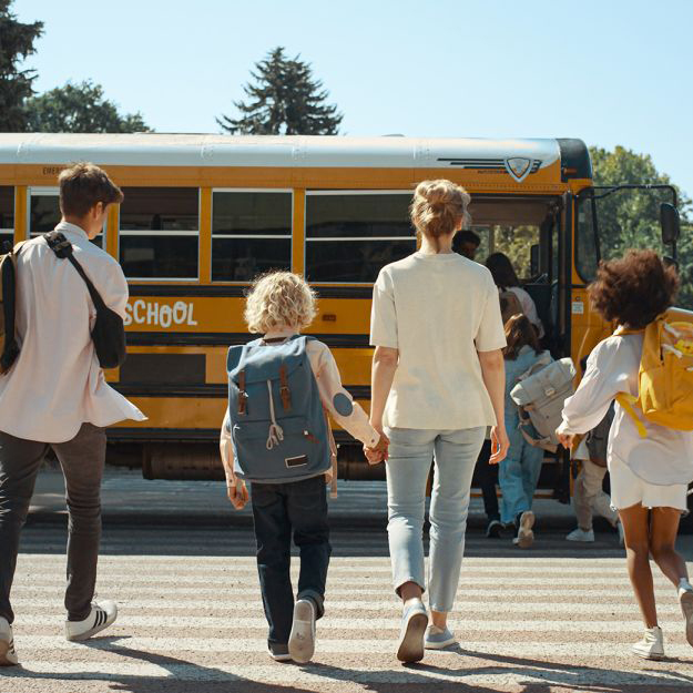 A group of children and an adult walk toward a yellow school bus at a crosswalk on a sunny day.